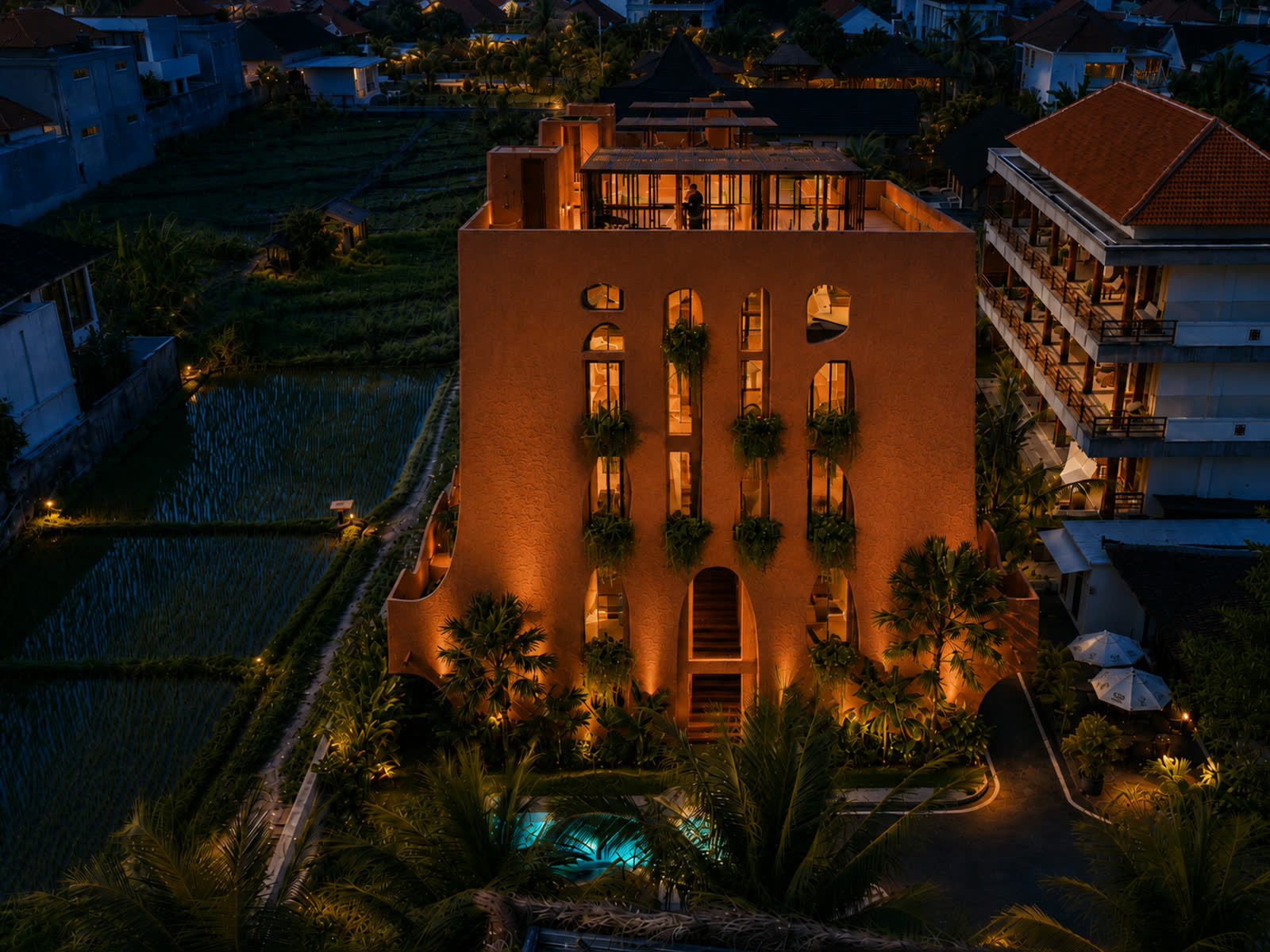 Night aerial view of Ananta Canggu's terracotta building beside rice paddies, with neighbouring rooftops and a lit rooftop terrace